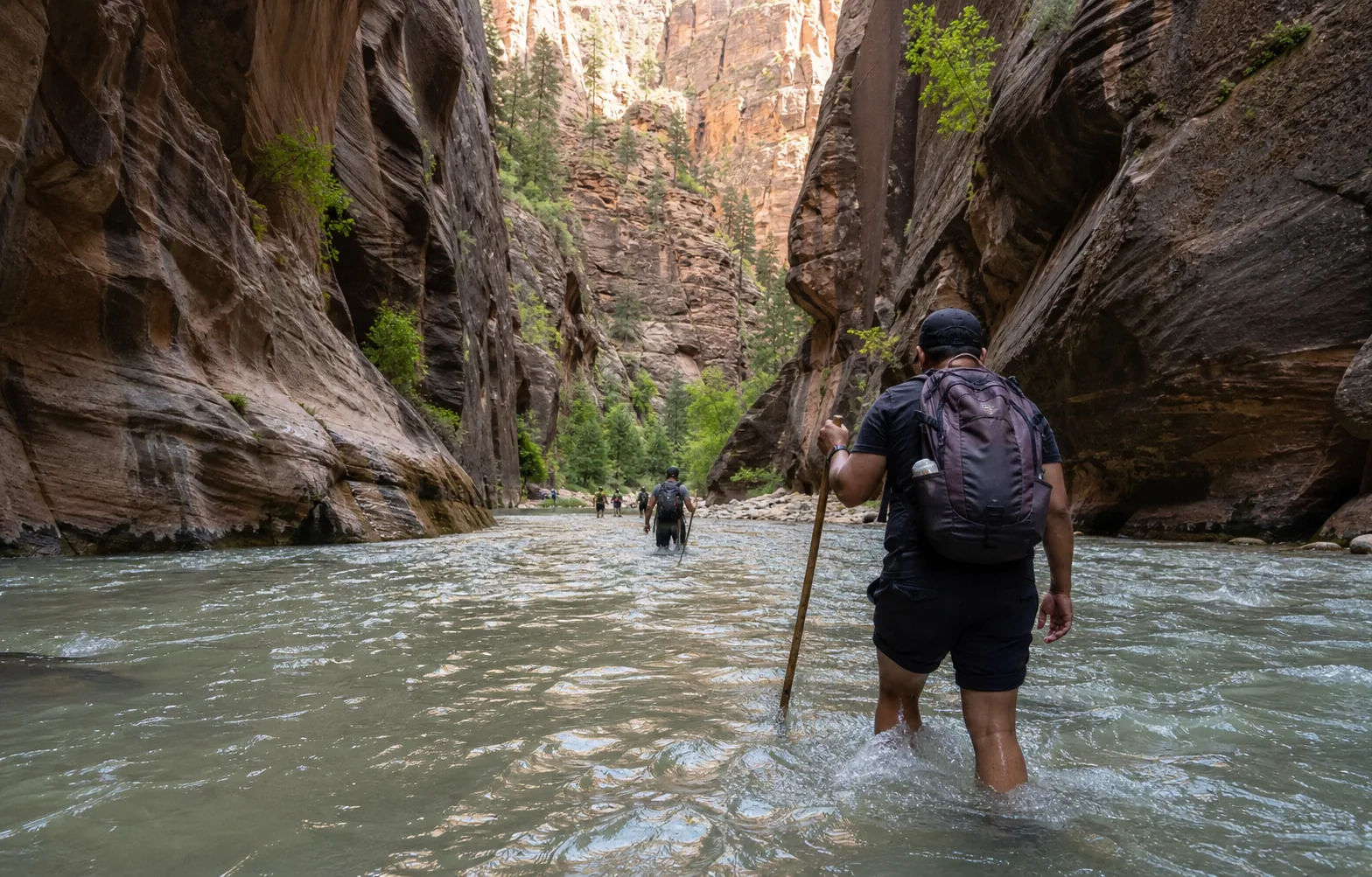 Hikers in closed-toe footwear moving carefully through the Virgin River between tall canyon walls.