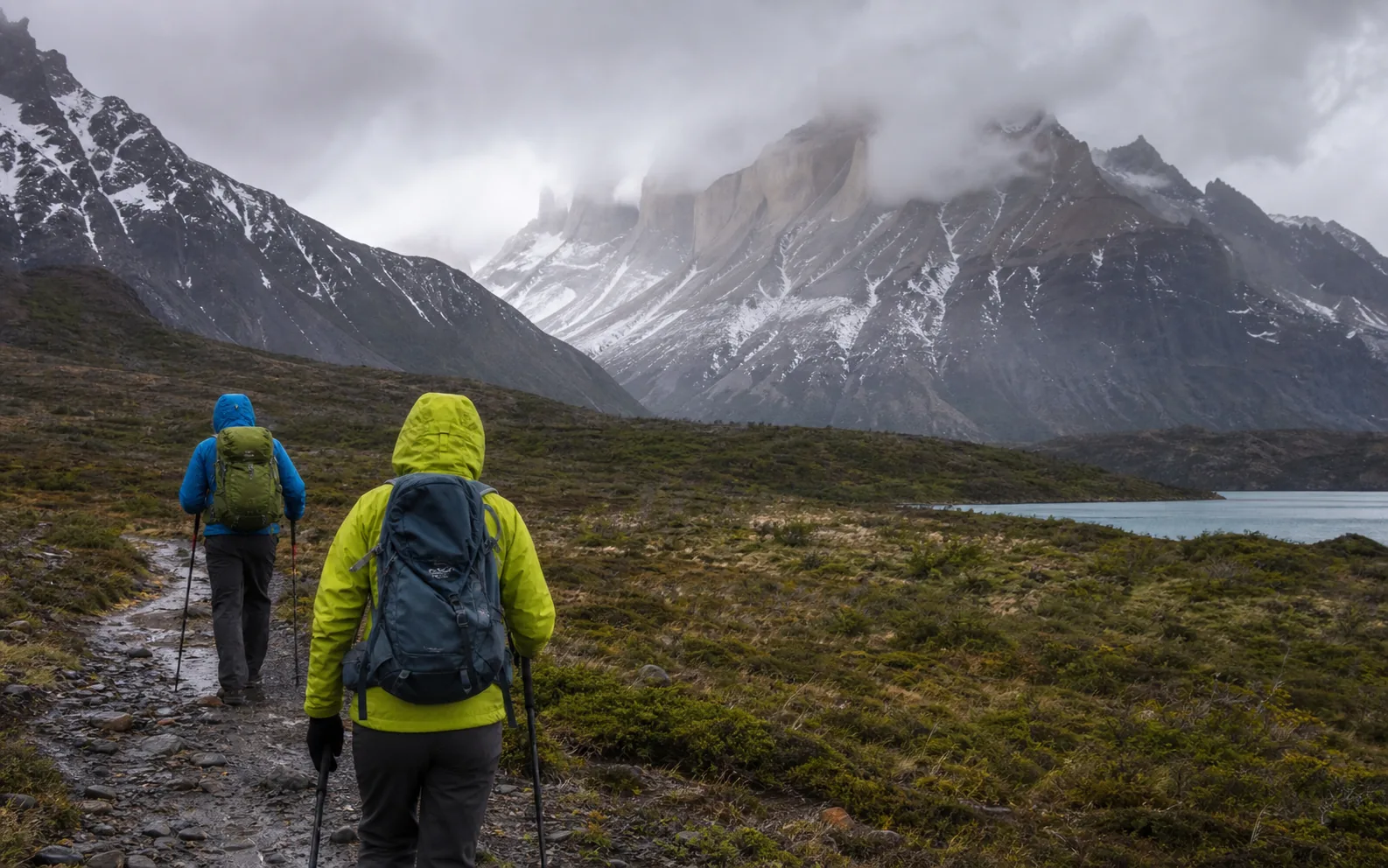 A trekker tightening a rain shell and backpack straps on the windy W Trek in Patagonia.